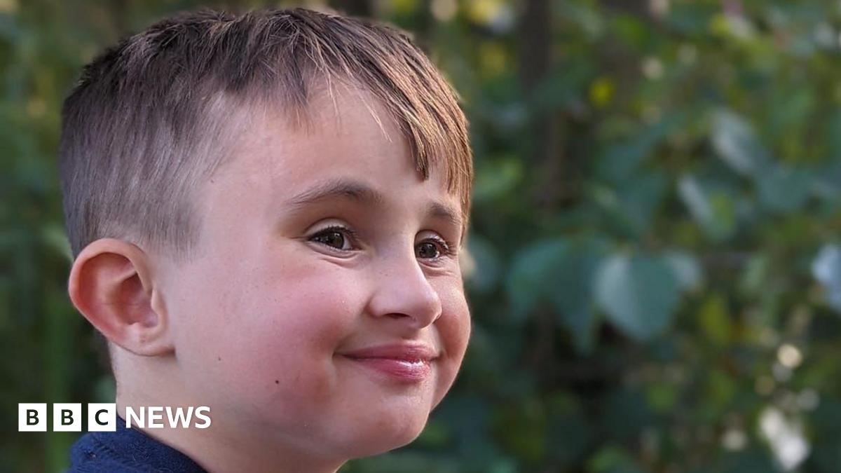 A close-up image of a young boy's face. He is looking away from the camera but smiling. There are green leaves from a tree in the background.