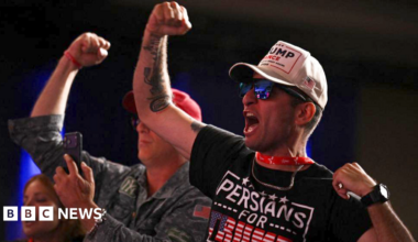 Matt Gaetz delivers remarks at a podium, looking to the right of the picture. He has his right arm outstretched and wears a dark blue suit and white shirt. He appears to be on stage under strong spotlights.