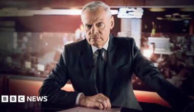Clunes as Edwards. He wears a white shirt, with black suit jacket and tie. his right hand is rested on the desk in front of him, with his left reaching out