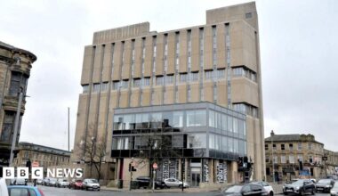 A large, multi‑storey concrete building on a street corner in an urban area. The building has a distinctive, Brutalist-style design, with tall vertical concrete panels running down the front.