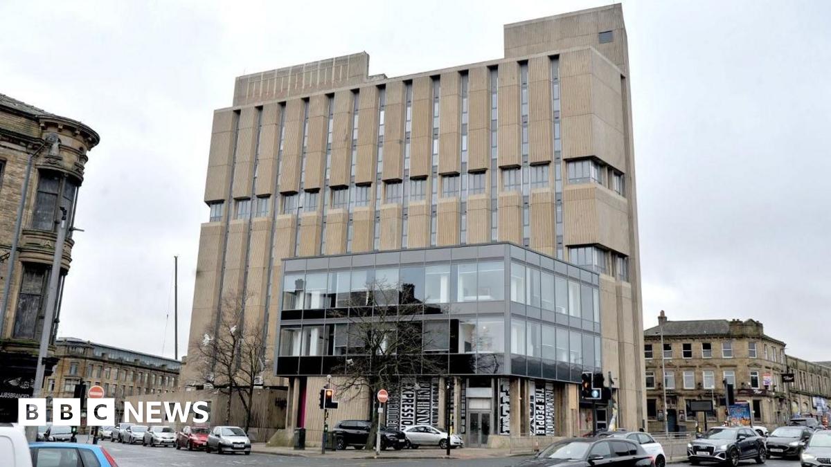 A large, multi‑storey concrete building on a street corner in an urban area. The building has a distinctive, Brutalist-style design, with tall vertical concrete panels running down the front.