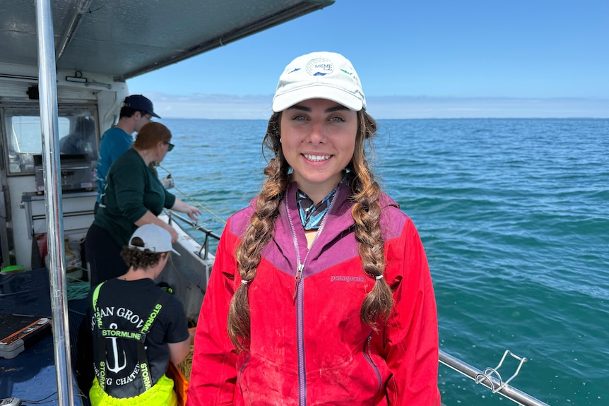girl with plaited hair wearing cap standing inside a boat wearing a red spray jacket.