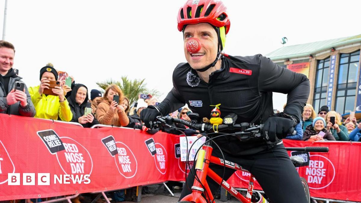 Radio 1 DJ Greg James is on his red bike on the starting line runway. Either side is a metal barrier lined with people waving. He is wearing all black cycling outfit and a red helmet and a red nose.