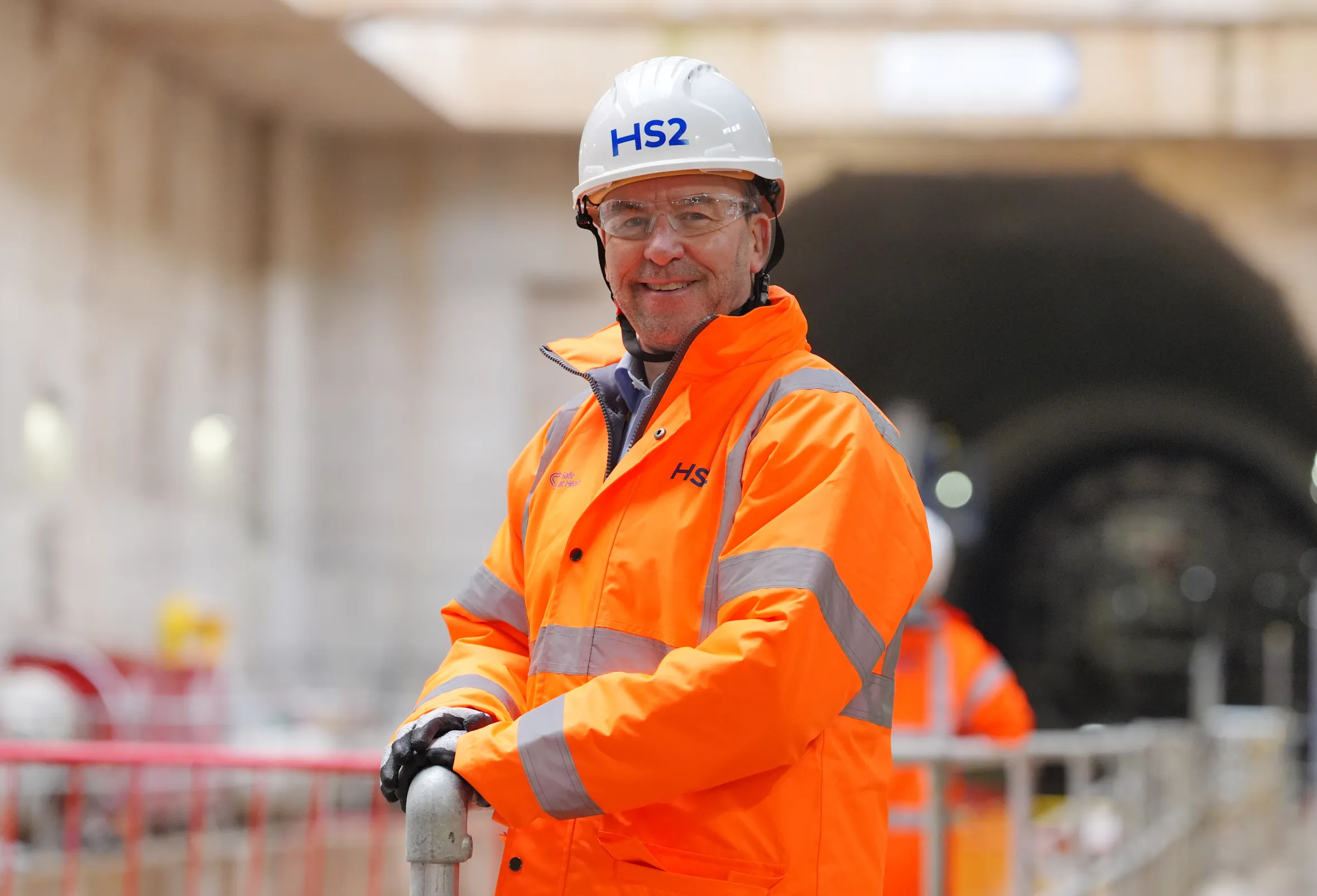 Mark Wild, HS2 CEO, wearing an HS2 hard hat, glasses, and an orange high-visibility jacket, smiling at the Old Oak Common station box site.