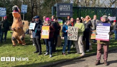 A group of people holding cardboard signs at a picket line with one person dressed like an orange dinosaur in an inflatable costume. They are standing in front of the blue Heriot Watt University Edinburgh campus sign