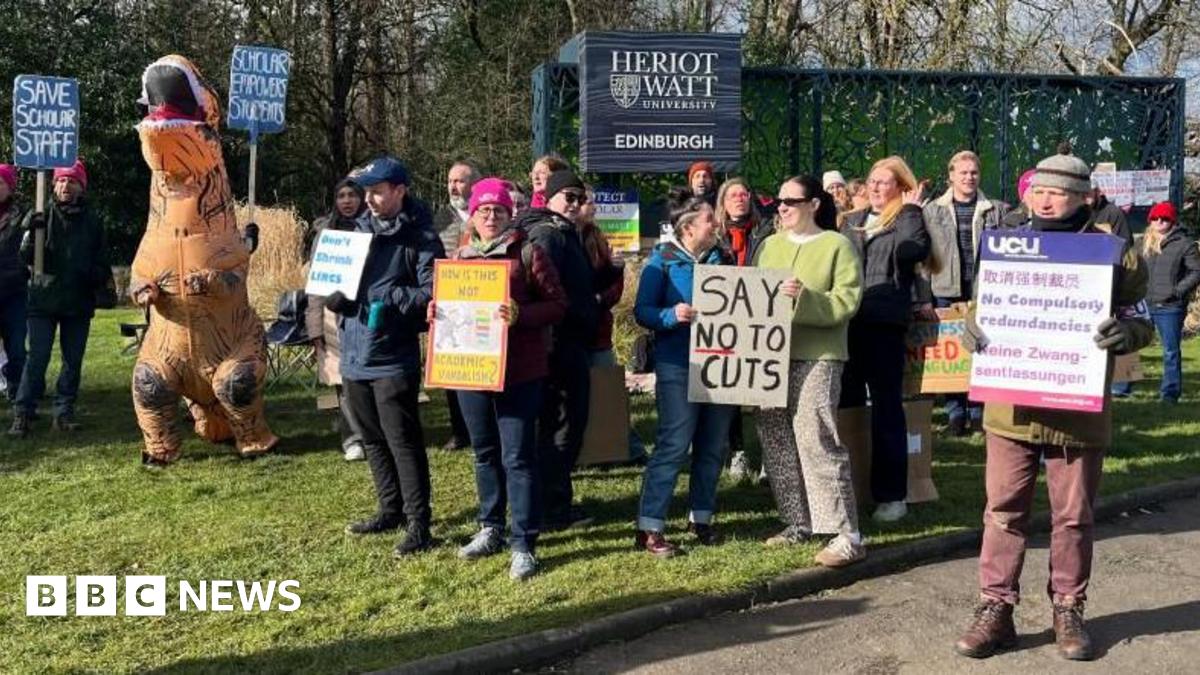 A group of people holding cardboard signs at a picket line with one person dressed like an orange dinosaur in an inflatable costume. They are standing in front of the blue Heriot Watt University Edinburgh campus sign