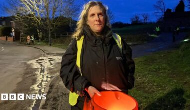 A woman with long hair, a black coat and a high-vis jacket, holds an orange bucket as she stands on a pavement next to a village road. Other volunteers can be seen behind her.