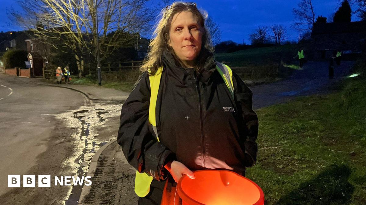 A woman with long hair, a black coat and a high-vis jacket, holds an orange bucket as she stands on a pavement next to a village road. Other volunteers can be seen behind her.