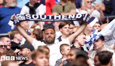 A bearded man in a white T-shirt holds a Southend United scarf above his head among a crowd of fans at Wembley Stadium