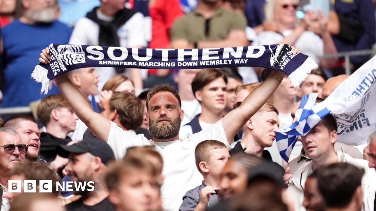 A bearded man in a white T-shirt holds a Southend United scarf above his head among a crowd of fans at Wembley Stadium