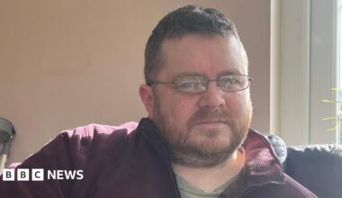 Mark has thin rectangular glasses and is wearing a burgandy fleece unzipped at the collar. He has short stubble and cropped brown hair.