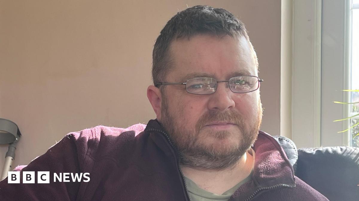 Mark has thin rectangular glasses and is wearing a burgandy fleece unzipped at the collar. He has short stubble and cropped brown hair.