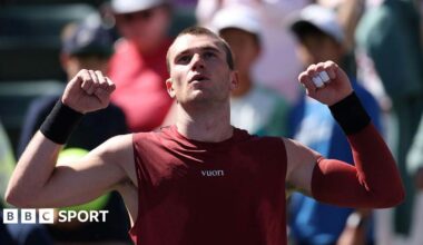 Jack Draper raises his arms to celebrate victory. He has short hair and is wearing a marron vest and a maroon sleeve on his left arm. Members of the crowd can be seen in the background.