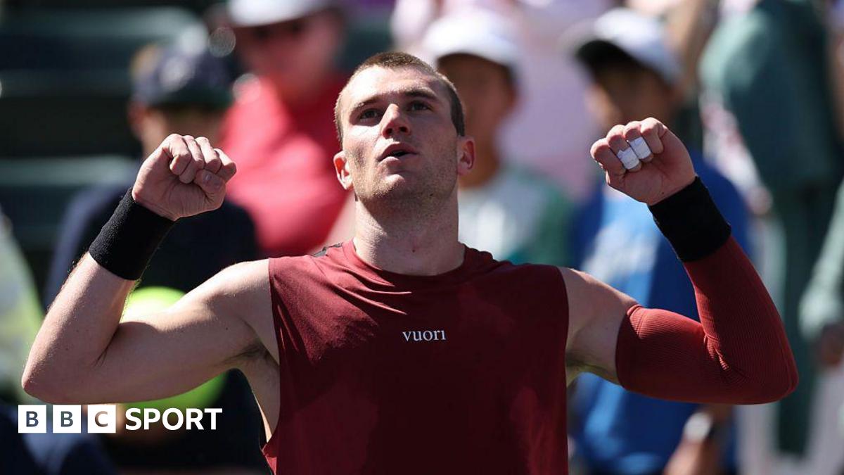 Jack Draper raises his arms to celebrate victory. He has short hair and is wearing a marron vest and a maroon sleeve on his left arm. Members of the crowd can be seen in the background.
