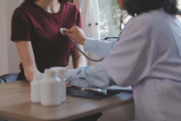 Doctor using a stethoscope to examine a patient with medicine bottles on the table behind in a clinical setting.