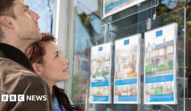 A woman with a blonde ponytail and a light grey trench coat holds a handbag and a paper bag while looking in a real estate window at real estate listings.