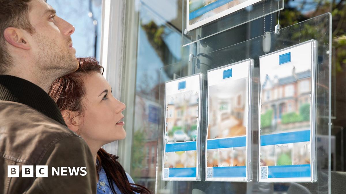 A woman with a blonde ponytail and a light grey trench coat holds a handbag and a paper bag while looking in a real estate window at real estate listings.