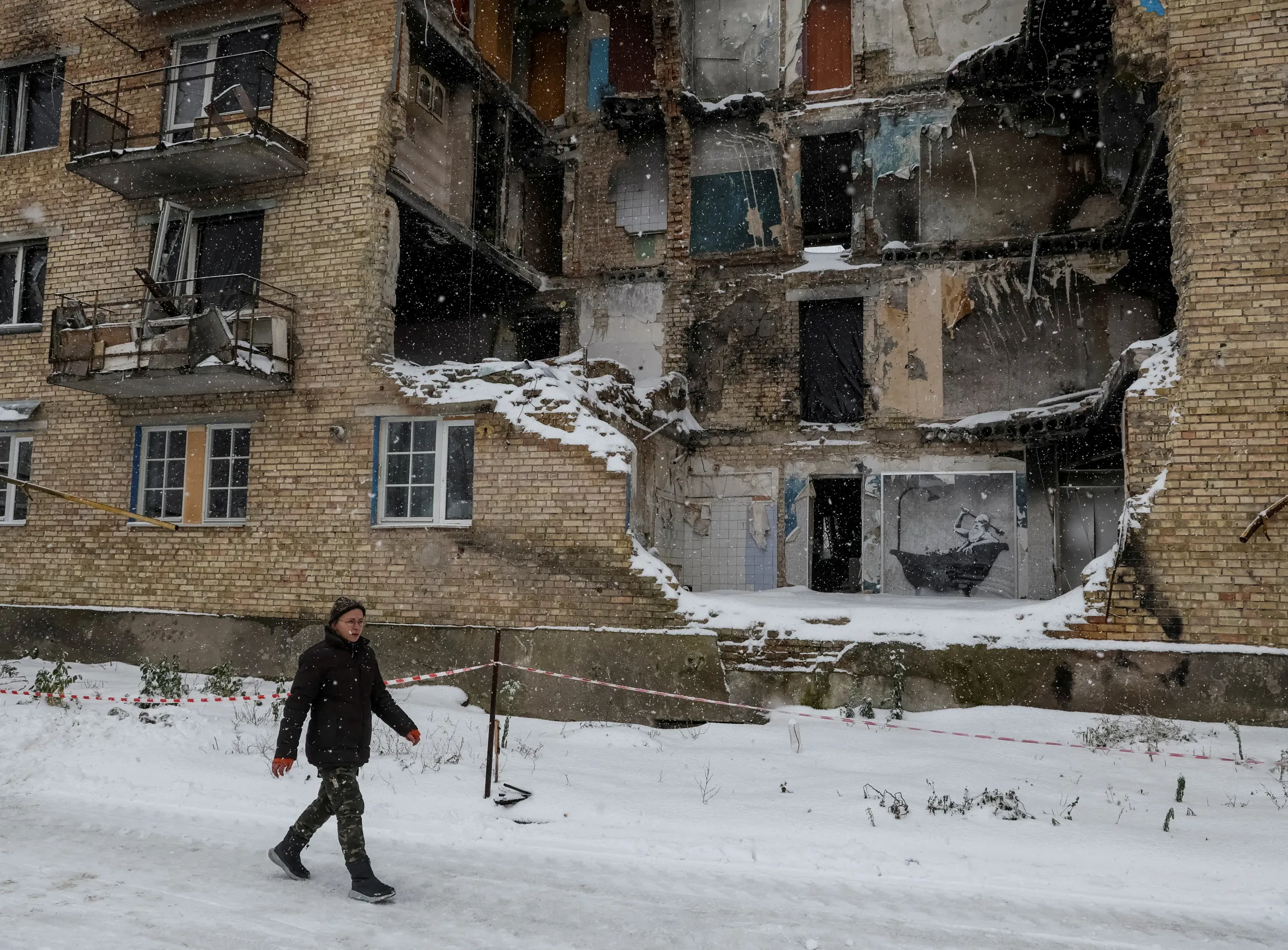 A person walks past a partially destroyed brick building with a Banksy graffiti artwork on its wall.