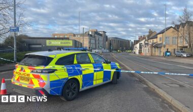 The rear of a police car is shown inside police tape on a road. In the distance, more police cars and officers can be seen