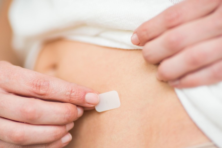 Close-up of torso of white woman. Focus is on her hand applying a small, white patch to the skin.