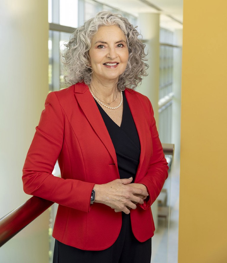 White woman with curly, grey hair and wearing a red jacket smiling to camera.