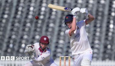 Rocky Flintoff driving a red cricket ball for Lancashire with his bat held high. He is wearing cricket whites and a dark blue Lancashire helmet with the Red Rose insignia on the front.