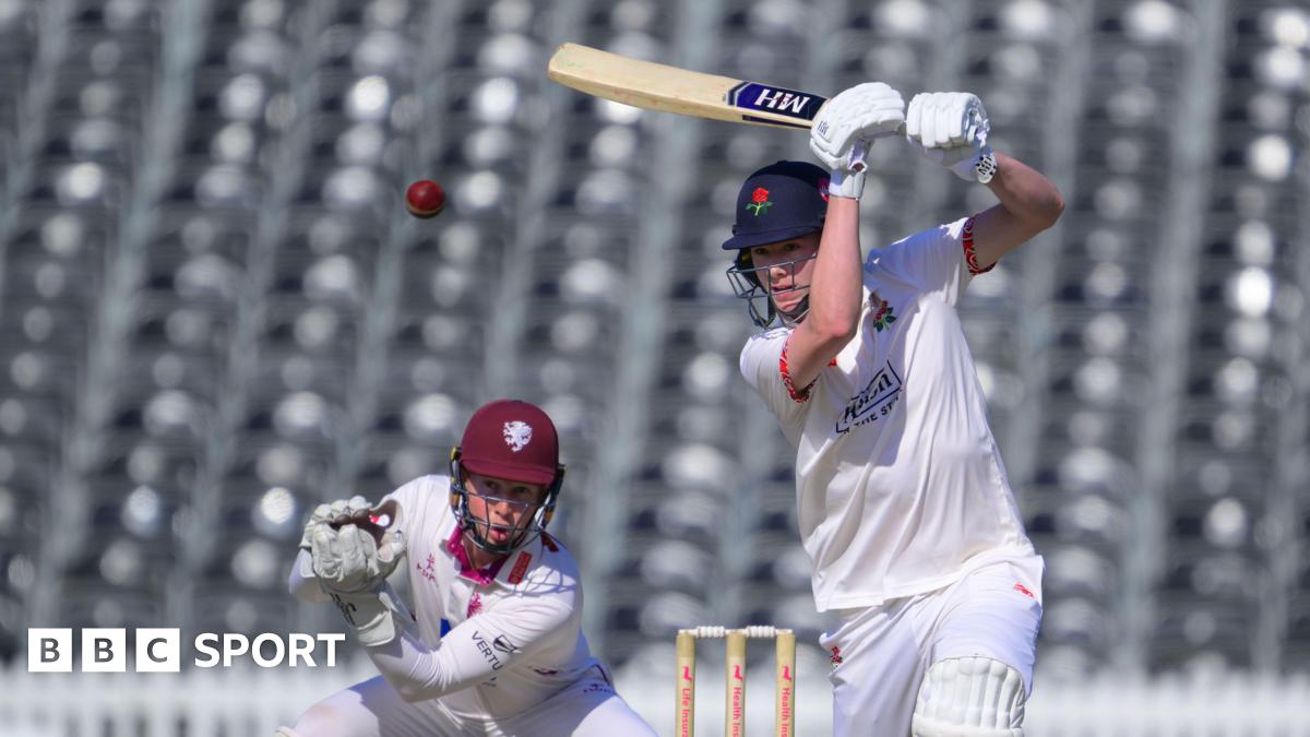 Rocky Flintoff driving a red cricket ball for Lancashire with his bat held high. He is wearing cricket whites and a dark blue Lancashire helmet with the Red Rose insignia on the front.