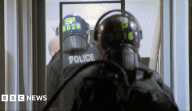 Two police officers wearing black tactical clothing and helmets with yellow writing, carrying riot shields.