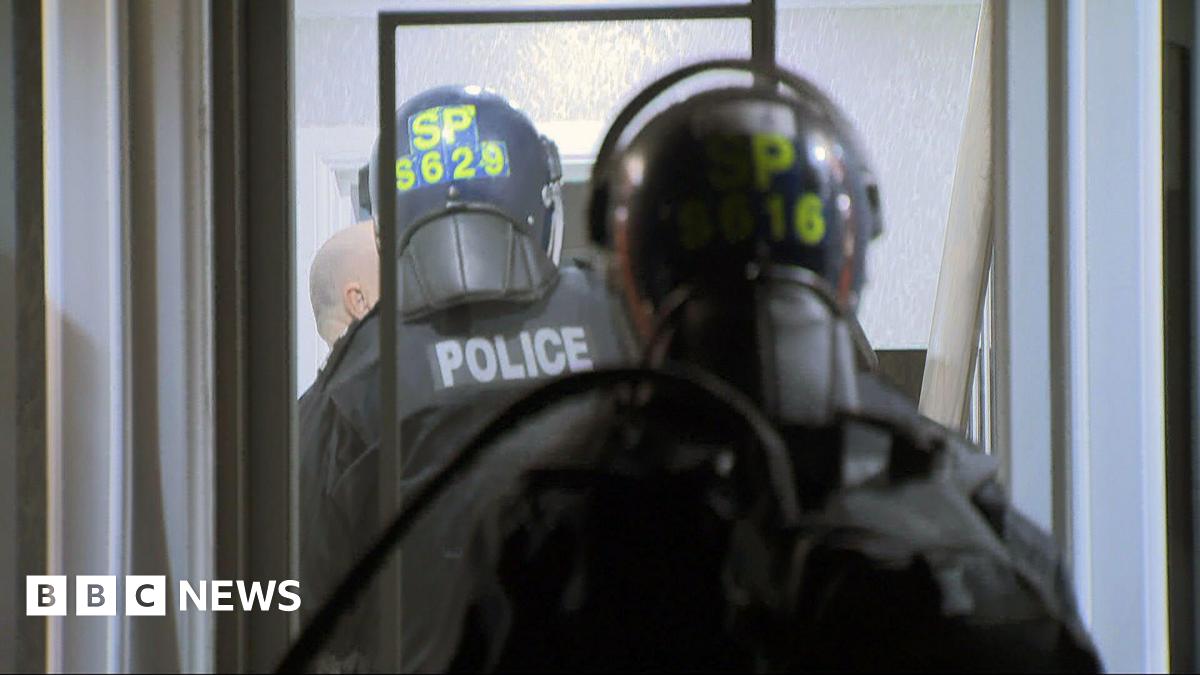 Two police officers wearing black tactical clothing and helmets with yellow writing, carrying riot shields.