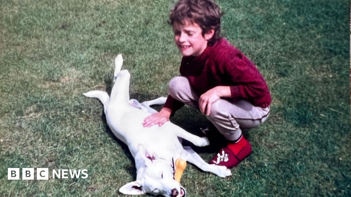 A grainy film photograph taken in the 1980s of a young boy playing on the grass with a white dog, who has a rubber bone in its mouth. The boy has short curly dirty blond hair and is wearing a red jumper, red slippers, and grey jogging bottoms. His eyes are closed and he is smiling.