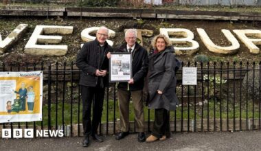 Horace Pickett posing for a picture standing beside two people outside the Westbury train station. He is holding an A3 frame in his hands, showing photos from his time on the team.