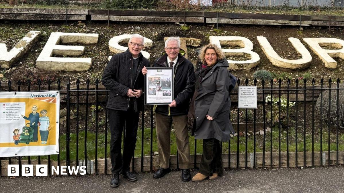 Horace Pickett posing for a picture standing beside two people outside the Westbury train station. He is holding an A3 frame in his hands, showing photos from his time on the team.