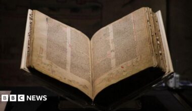A medieval book open on a stand on a table, which a black background. The book has yellowing pages covered with five columns of densely written text and drawings around the edges.