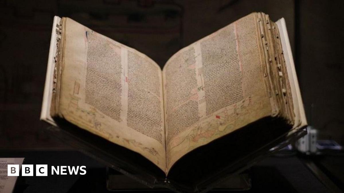 A medieval book open on a stand on a table, which a black background. The book has yellowing pages covered with five columns of densely written text and drawings around the edges.