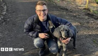 a young man wearing glasses wearing a parka jacket and jeans with his arm round a black dog. He is kneeling on a muddy path.