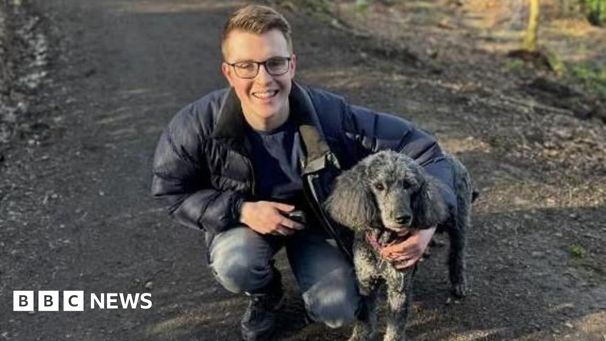 a young man wearing glasses wearing a parka jacket and jeans with his arm round a black dog. He is kneeling on a muddy path.