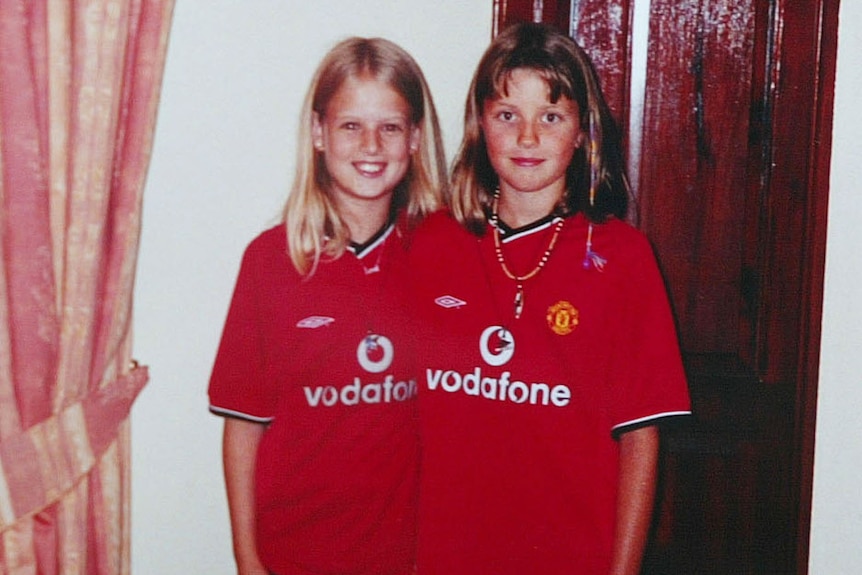 Two girls, in red Manchester United Football Club shirts, stand by a brown door. A clock on the wall shows 5.04pm.
