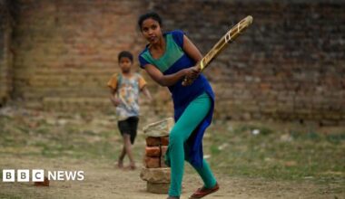 A woman with dark hair and a multiple colour jumper siting on a green sofa inside her living room.
