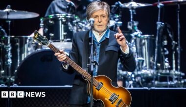Paul McCartney on stage wearing a blue jacket, blue shirt and with a guitar. He is pointing one finger in the hair.