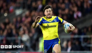 A photo of Matty Ashton running with the ball in his Warrington Wolves kit with the crowd in the background and an opposing player on the floor behind him
