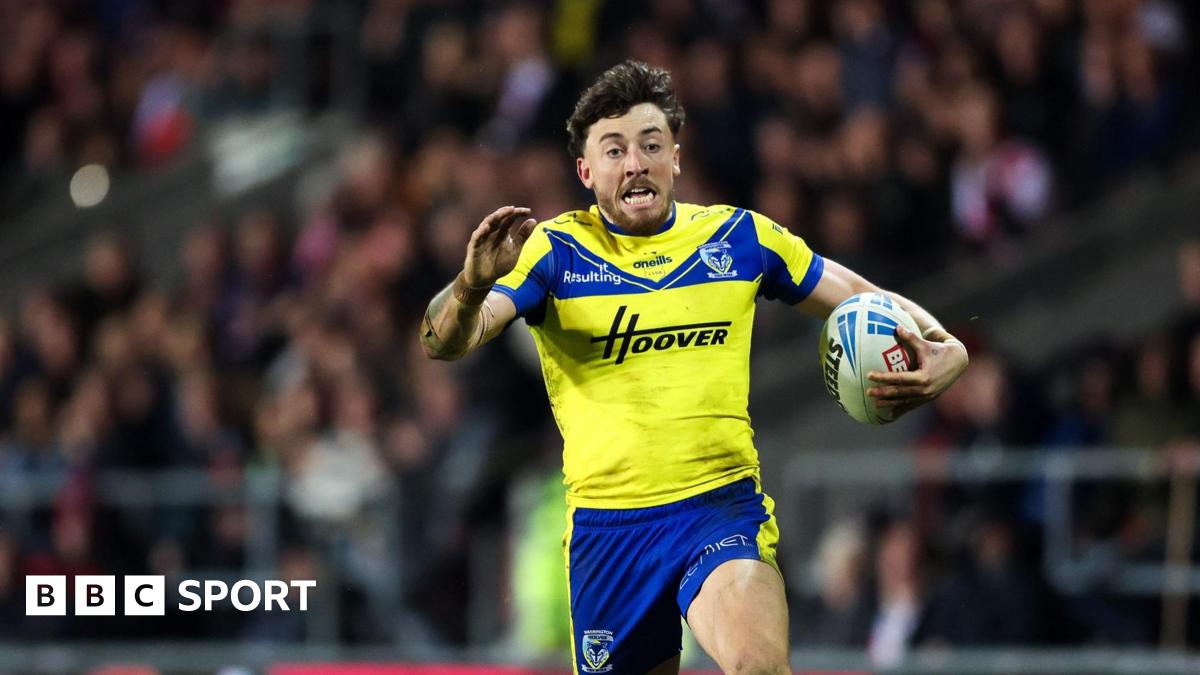 A photo of Matty Ashton running with the ball in his Warrington Wolves kit with the crowd in the background and an opposing player on the floor behind him