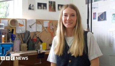 The image shows Iona Hall wearing a white t-shirt underneath a denim pinafore, with silver chains layered around her neck. She has long blonde hair and is smiling at the camera, sitting at a desk full of tools used to make jewellery.
