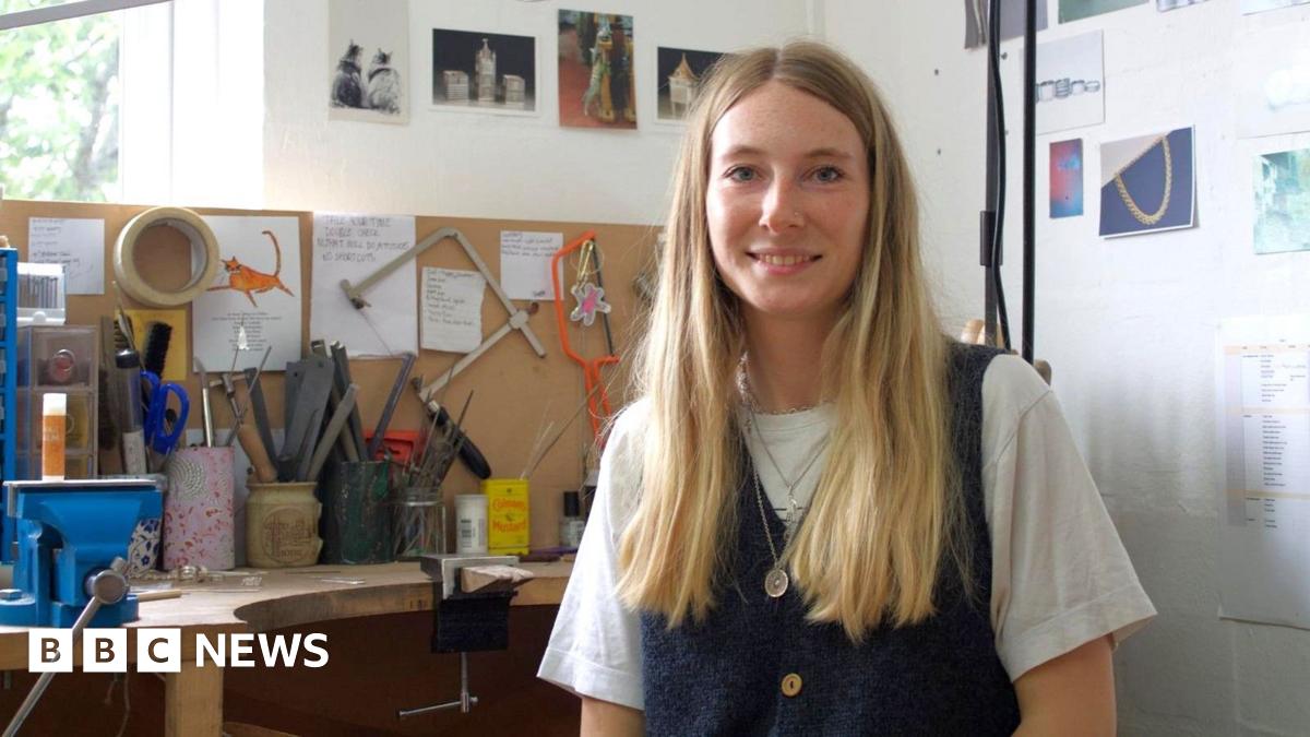 The image shows Iona Hall wearing a white t-shirt underneath a denim pinafore, with silver chains layered around her neck. She has long blonde hair and is smiling at the camera, sitting at a desk full of tools used to make jewellery.