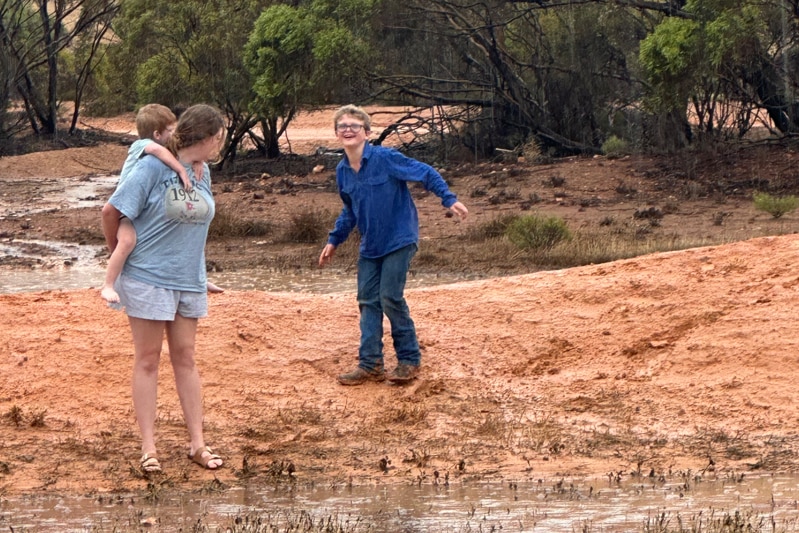 A young woman with a little boy on her back looks at a laughing teenage boy as they stand on a muddy paddock.