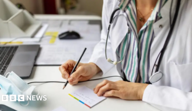 A person in a white coat with with a pencil writing on a piece of paper in a GP surgery setting