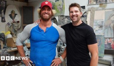 Nick Elphick wears a blue and grey long-sleeved top and red baseball hat. He is standing next to Darryn Frost, who has his arm around him. He is wearing a black T-shirt. They are both smiling at the camera and standing in an art studio, with paintings and small sculptures in the background.
