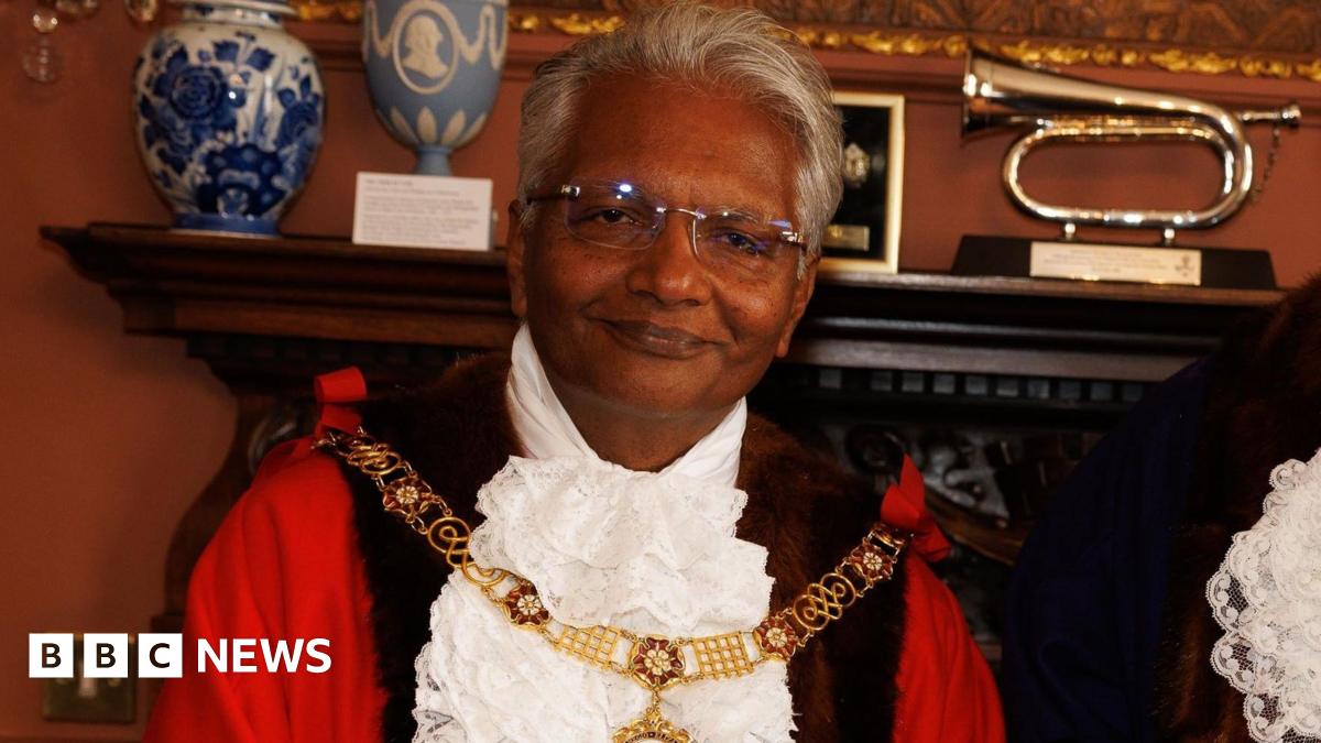 Wearing his mayoral robes and gold chain, Bharat Pankhania smiles at the camera. Behind him two vases and a musical instrument can be seen on a mantelpiece.