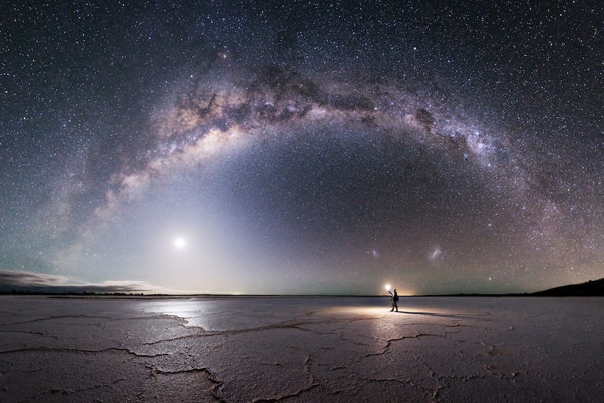 Michael stands small holding a small light under the milky way on a dry cracked lake.