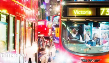 A red double-decker London bus stuck in traffic on Oxford Street at night time. There are street lights lighting up the vehicles, and the bus's bright headlights are shining at the camera. The bus is travelling to Victoria. A bus driver is sitting in the cabin at the front behind the wheel, and there are people stood on board the bus. A queue of London buses and black taxis are visible to the left of the bus.