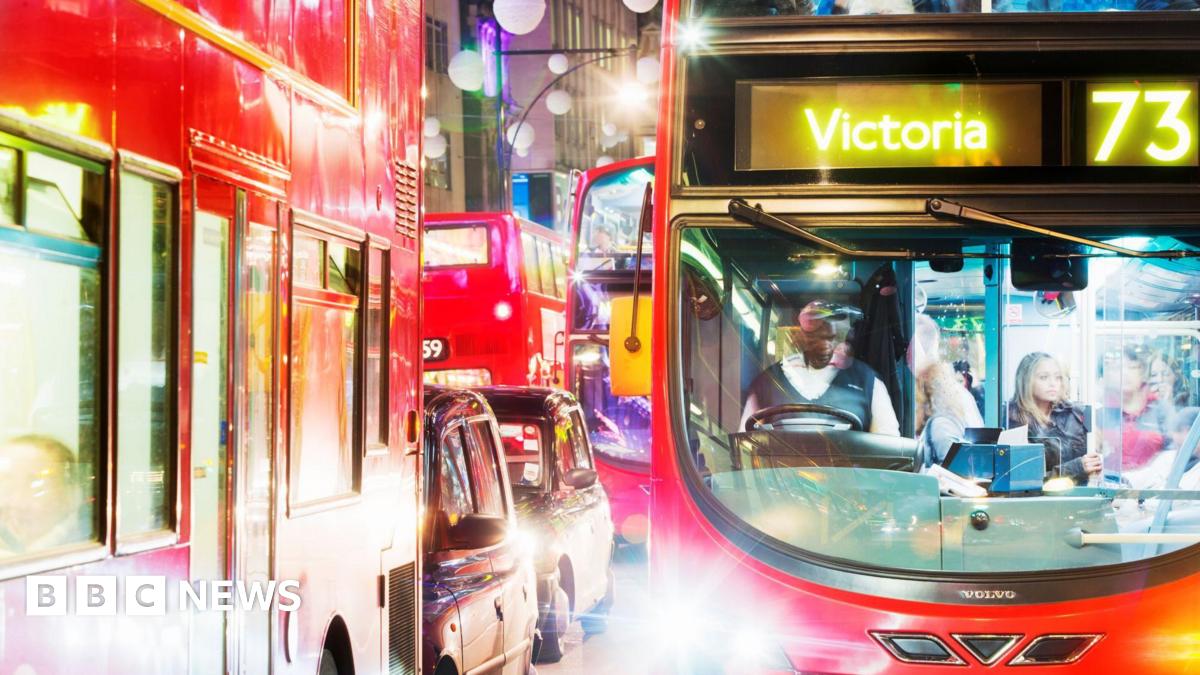 A red double-decker London bus stuck in traffic on Oxford Street at night time. There are street lights lighting up the vehicles, and the bus's bright headlights are shining at the camera. The bus is travelling to Victoria. A bus driver is sitting in the cabin at the front behind the wheel, and there are people stood on board the bus. A queue of London buses and black taxis are visible to the left of the bus.